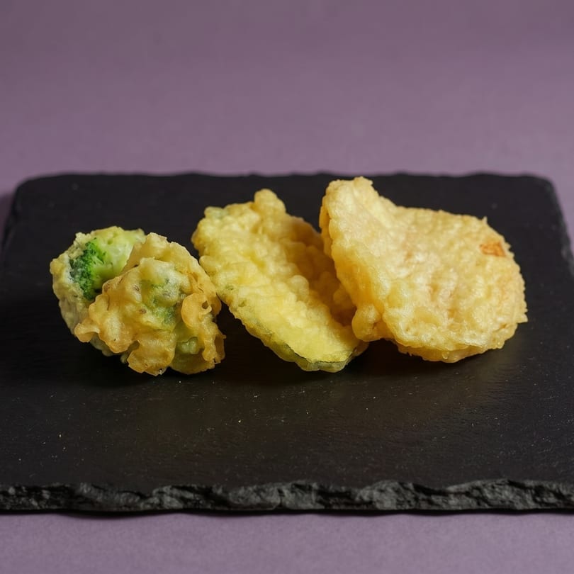 Three pieces of vegetable tempura, including broccoli and zucchini, on a dark slate board.