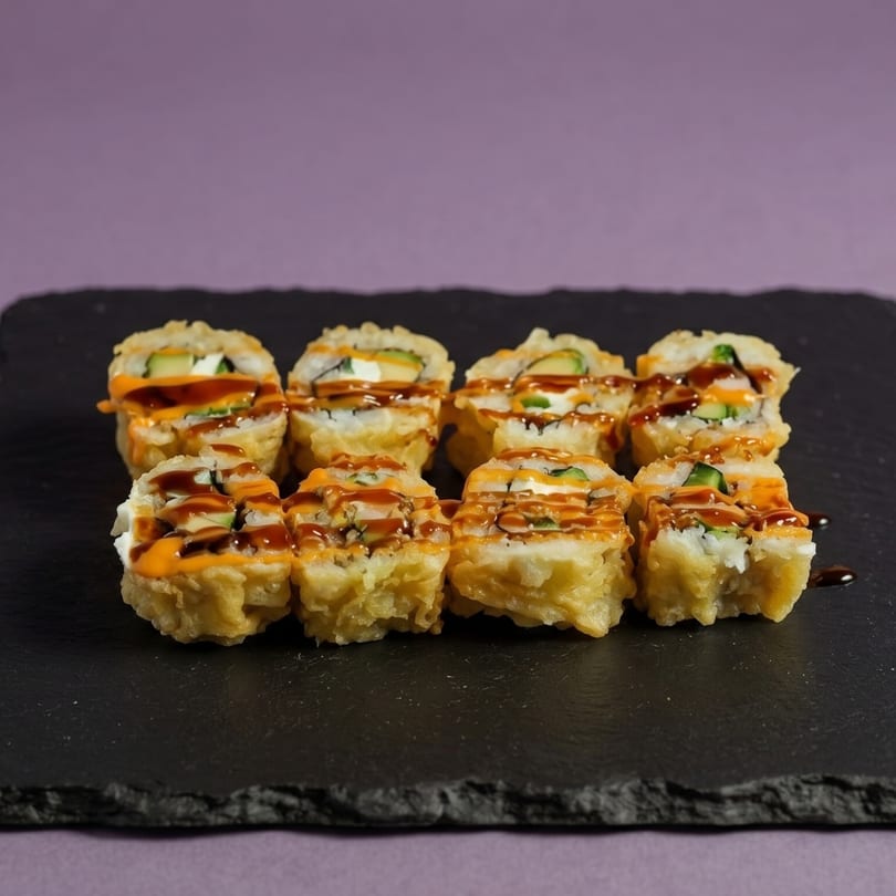 Eight pieces of deep-fried sushi rolls with orange and brown sauce on a black slate.
