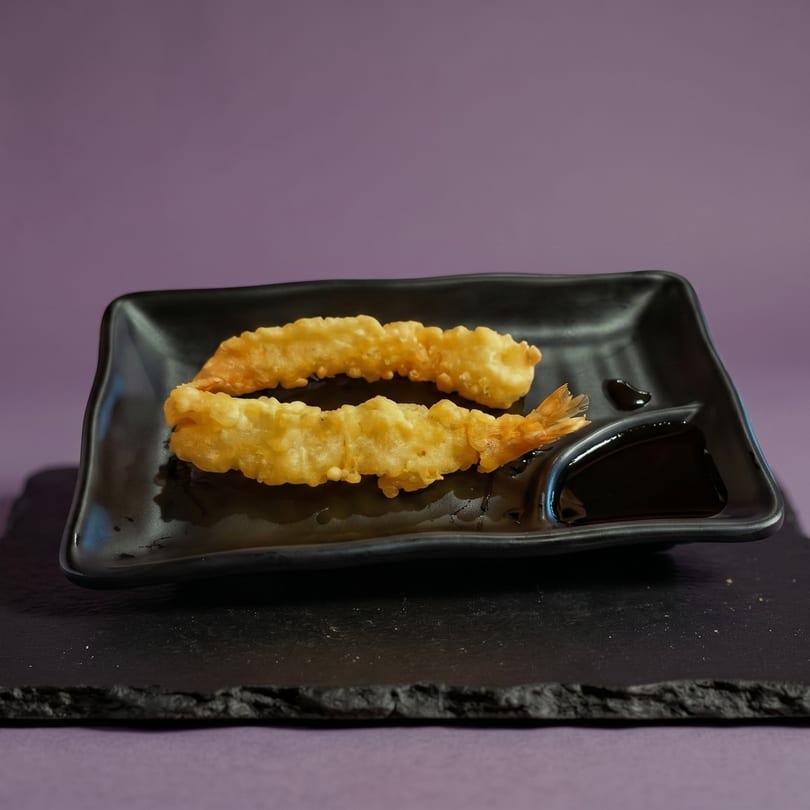 Two golden tempura shrimp with a side of dark dipping sauce on a black plate.